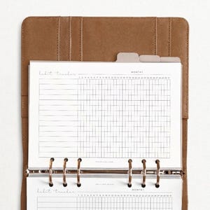 Può includere: Un raccoglitore in pelle marrone contenente pagine bianche per il monitoraggio delle abitudini con una griglia. Le pagine riportano il testo "habit tracker" e "month" con colonne numerate da 1 a 31 e sei anelli metallici di fissaggio.