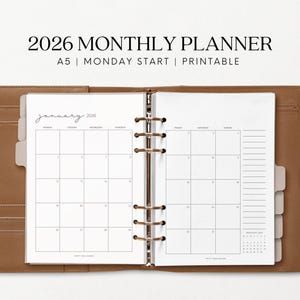 May include: A brown leather A5 planner open to a monthly calendar page for January 2026. The planner has a ring binder mechanism and includes the text "2026 MONTHLY PLANNER" and "A5 | MONDAY START | Printable".