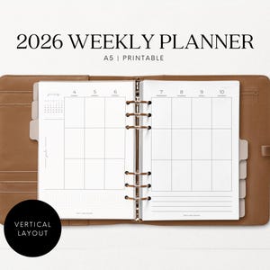 May include: Open A5 weekly planner for 2026, featuring a vertical layout. The planner is in a brown leather-like cover with a ring binder. The pages are white with gray grid lines and the text "2026 WEEKLY PLANNER".