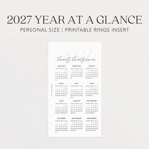 May include: A white printable calendar insert for 2027, featuring a year-at-a-glance layout. The design includes the text "2027 YEAR AT A GLANCE" and "PERSONAL SIZE | PRINTABLE RINGS INSERT". The calendar displays the months and dates in a clear, readable format.