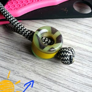May include: A close-up of a lanyard bead with a camouflage pattern in green, brown, and yellow. The bead is attached to a black and white rope. A pink and black knife is in the background.