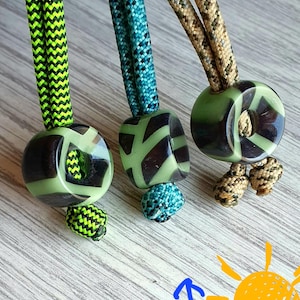 Puede incluir: Tres cordones decorativos con cuentas estampadas. Cada cordón tiene una cuerda de diferente color: verde neón, verde azulado y beige. Las cuentas son verdes y negras con un diseño geométrico. Los cordones están sobre una superficie de madera clara.