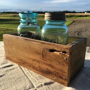 May include: A rustic wooden box holds two vintage blue glass mason jars with metal lids. The jars are clear, and the box is made of weathered wood with visible nail heads. The scene is set outdoors with a soft, natural light.