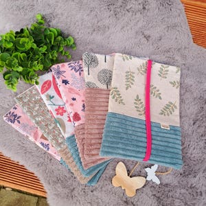 Könnte beinhalten: Eine Sammlung von Stoff-Buchhüllen mit verschiedenen Blumen- und Baummustern. Die Hüllen zeigen eine Mischung aus rosa, beige und blauen Stoffen, einige mit Cordstrukturen. Ein leuchtend rosa Band ist an einer Hülle befestigt. Ein Holzschmetterling und -stern sind davor platziert.