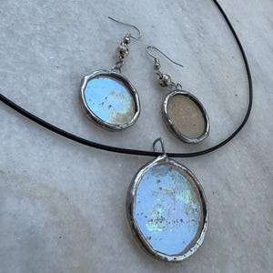 Könnte beinhalten: Ein Schmuckset mit Anhänger und passenden Ohrringen. Der Anhänger und die Ohrringe haben eine ovale Form mit einem silberfarbenen Metallrand. Der Anhänger hängt an einer schwarzen Kordel. Die Ohrringe haben einen Hakenverschluss.