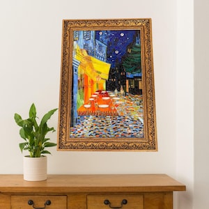 May include: A framed mosaic artwork depicting a nighttime street scene with a cafe. The artwork features a starry sky, buildings, and a cafe with tables and chairs. The frame is gold-colored.
