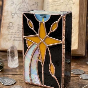 May include: A stained glass tarot card box with a black background and a yellow star design. The box has copper-colored edging and contains tarot cards. The top card reads "XVII". The box is on a wooden surface.