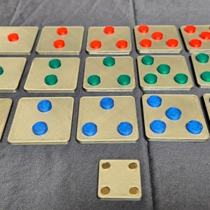 May include: A collection of square game tiles with gold-colored bases and colored circular markers. The markers are red, green, and blue. The tiles are arranged on a gray surface.