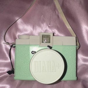 May include: A mint green and cream-colored Diana+ film camera with a textured body, a round lens cap featuring the embossed text DIANA+, a top-mounted viewfinder, and an attached off-white carrying strap extending from the sides of the camera.