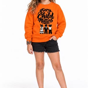 May include: A bright orange long-sleeve sweatshirt featuring the black text Every Child Matters above a graphic of three children holding hands. The sweatshirt is paired with black denim shorts and white sneakers, creating a casual outfit.