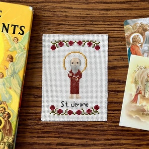May include: A white cross-stitch embroidery of Saint Jerome, a bearded man in a red robe, with a halo above his head. The embroidery is framed with a green vine and red roses.