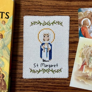 Könnte beinhalten: Ein Kreuzstich-Bild von St. Margaret mit Heiligenschein und blauen Gewändern, darunter der Name. Das Bild ist von grünen floralen Akzenten umgeben. Religiöse Karten und ein Buch mit dem Titel "More Saints" sind ebenfalls zu sehen.