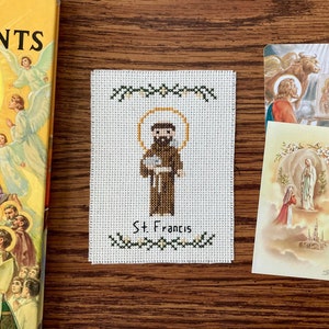 May include: A white cross-stitch embroidery of Saint Francis of Assisi. The embroidery is framed with a green border and features a brown figure of Saint Francis holding a bird in his hand. The text "St. Francis" is written below the figure.