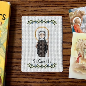 May include: A white cross-stitch embroidery of Saint Colette, a Catholic saint, with a halo and a brown robe. The embroidery is framed with a green vine and blue flowers. The text "St. Colette" is written below the image.