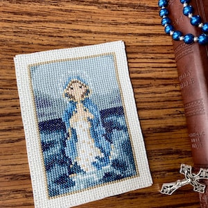 Op de afbeelding: Een kruissteekborduursel van de Maagd Maria die op een blauwe en witte golf staat. Het borduursel is ingelijst in een wit frame met een gouden rand.