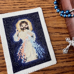 Puede incluir: Un bordado de punto de cruz de Jesucristo con un fondo azul y un halo blanco. El bordado está enmarcado con un borde blanco.