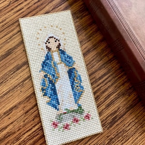 Può includere: Un segnalibro a punto croce che raffigura la Vergine Maria con una veste blu e un abito bianco, in piedi su una base verde con rose rosse. Lo sfondo è di colore crema con un bordo dorato.