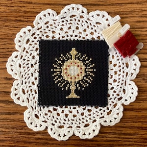 May include: A black cross stitch embroidery on white fabric with a gold and red design of a monstrance. The embroidery is on a white doily and there are three spools of embroidery floss in the top right corner.