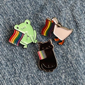 Może przedstawiać: Trzy szpilki emaliowane przedstawiające zieloną żabę, czarnego kota i białą gęś. Każde zwierzę trzyma tęczową flagę dumy.