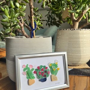 Puede incluir: Una obra de arte enmarcada que presenta tres plantas en maceta hechas con piezas de vidrio de colores. Las plantas están en macetas de diferentes colores y tienen flores rosas, rojas y blancas. La obra está firmada "LK Wolf" en la esquina inferior derecha.