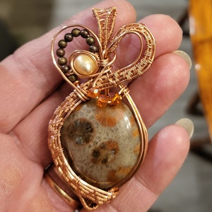 May include: A copper wire wrapped pendant featuring a brown and white fossilized coral stone with orange accents. The pendant is adorned with a pearl and brown beads.