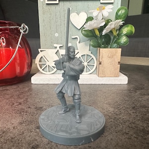 Puede incluir: Una figurita de plástico gris de un personaje de Star Wars sosteniendo un sable de luz. La figurita está de pie sobre una base gris con un diseño texturizado.