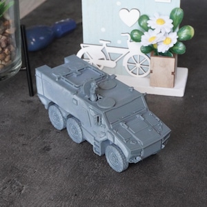 May include: A grey model of an armoured vehicle with six wheels, featuring detailed hatches and a gun turret. This miniature military vehicle is a detailed replica, showcasing the design of a modern armoured personnel carrier. The model is displayed on a dark surface.