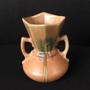 May include: A small, peach-colored ceramic vase with a flared, geometric top and two curved handles. The vase has a rounded base and a vertical green stripe down the front. The interior is a lighter peach color.