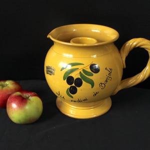 May include: A yellow ceramic jug with a twisted handle and a round base. The jug has a spout and a circular opening at the top. It is decorated with a branch of olives and the text "de Barjols". Two red and green apples are next to the jug.