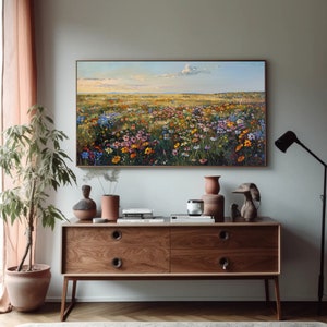 May include: A framed landscape painting depicts a vibrant field of wildflowers under a blue sky with clouds. The artwork hangs above a wooden cabinet with decorative items, including vases and a lamp. The color palette includes greens, yellows, blues, and pinks.