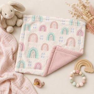May include: A soft, cream-colored baby blanket featuring a repeating pattern of pastel rainbows and blue raindrops, with a solid dusty pink plush underside. The blanket is displayed alongside a beige plush bunny, a pink pacifier, and a wooden rainbow teether.