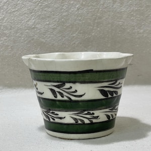 May include: A small, white ceramic pot with a scalloped rim. The pot features horizontal bands of green and white, with black leaf designs. The pot is likely used for planting small flowers or herbs.