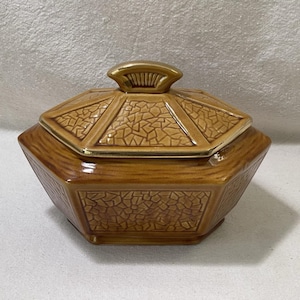 May include: A brown ceramic lidded dish with a geometric design. The lid has a gold-colored handle and trim. The dish has a textured, mosaic-like pattern on the lid and sides, and is octagonal in shape.