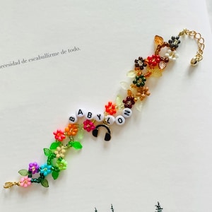 May include: A beaded bracelet featuring colorful flower-shaped beads in shades of pink, blue, green, orange, and red, interspersed with green leaf charms. It includes white letter beads spelling BABYLON and a small black headphone charm.