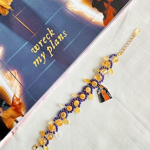 Puede incluir: Una pulsera dorada con cuentas moradas y amarillas, y un dije. La pulsera tiene la palabra "Willow" escrita con cuentas amarillas. La pulsera está al lado de un libro con las palabras "wreck my plans".