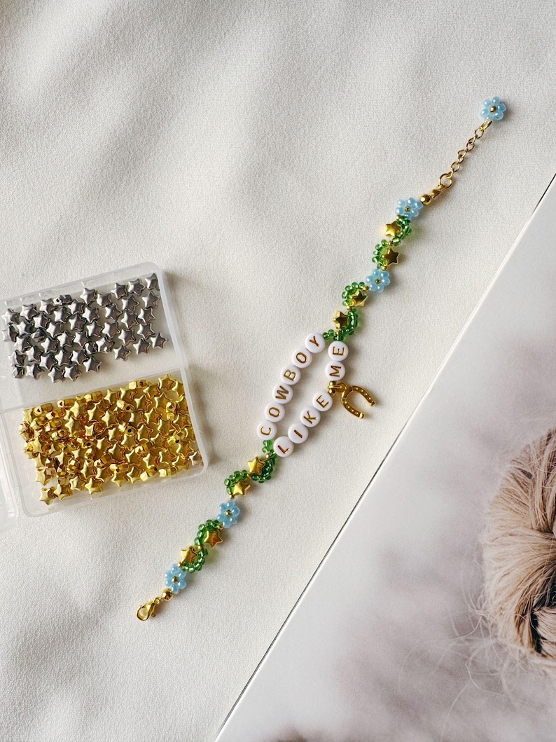 May include: A DIY bracelet kit with green and blue beads, gold stars, and white letter beads that spell "COWBOY LIKE ME". The kit also includes a horseshoe charm and two containers of silver and gold star beads.