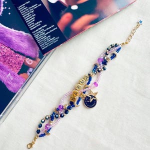 May include: A beaded bracelet with the word "KARMA" spelled out in gold letters. The bracelet features blue, purple, and clear beads, with a gold star charm and a blue and gold circular charm. A gold chain and clasp complete the design.
