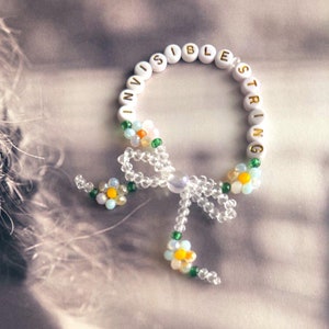 Puede incluir: Pulsera de cuentas blancas con las palabras "Invisible String" escritas en letras doradas. La pulsera tiene un lazo hecho de cuentas blancas y pequeños detalles de flores.