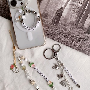 Puede incluir: Una colección de colgantes para teléfono y llaveros con la palabra "FOLKLORE" escrita con cuentas de letras blancas. Los colgantes presentan varios elementos decorativos, incluyendo estrellas, flores y objetos en miniatura. Un colgante está unido a una funda de teléfono.