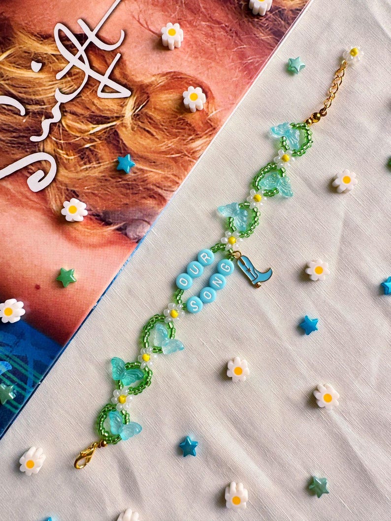 K&ouml;nnte beinhalten: Ein Perlenarmband mit einer goldenen Kette, das hellblaue Schmetterlingsanh&auml;nger, wei&szlig;e G&auml;nsebl&uuml;mchen-Akzente und hellblaue Buchstabenperlen mit der Aufschrift "OUR SONG" aufweist. Ein kleiner blauer Cowboy-Stiefel-Anh&auml;nger ist ebenfalls enthalten. Das Armband ist von dekorativen stern- und bl&uuml;tenf&ouml;rmigen Verzierungen umgeben.