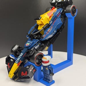 May include: A Lego model of a Formula 1 race car in navy blue, yellow, and red, with sponsor logos. The car is mounted on a blue stand, and a Lego figure of a driver in a blue racing suit and helmet stands beside it.