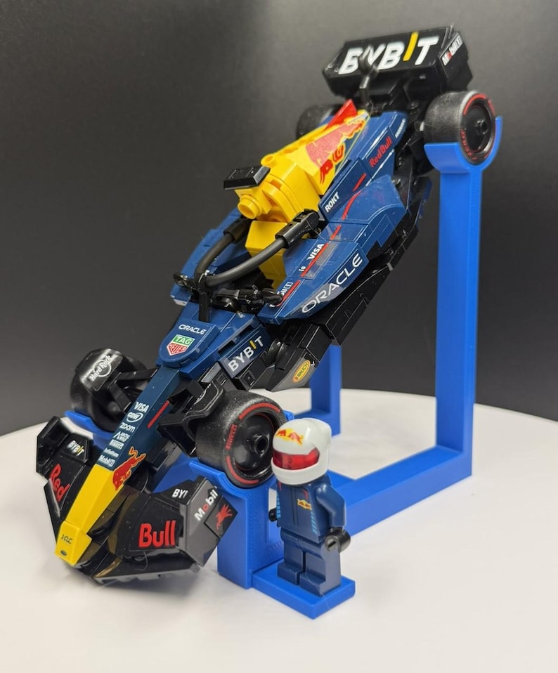 May include: A Lego model of a Formula 1 race car in navy blue, yellow, and red, with sponsor logos. The car is mounted on a blue stand, and a Lego figure of a driver in a blue racing suit and helmet stands beside it.