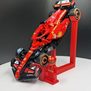 May include: A red and black Lego Formula 1 race car model with sponsor logos, displayed on a red stand. A red Lego figure of a driver stands next to the car. The car has black tires and yellow accents.