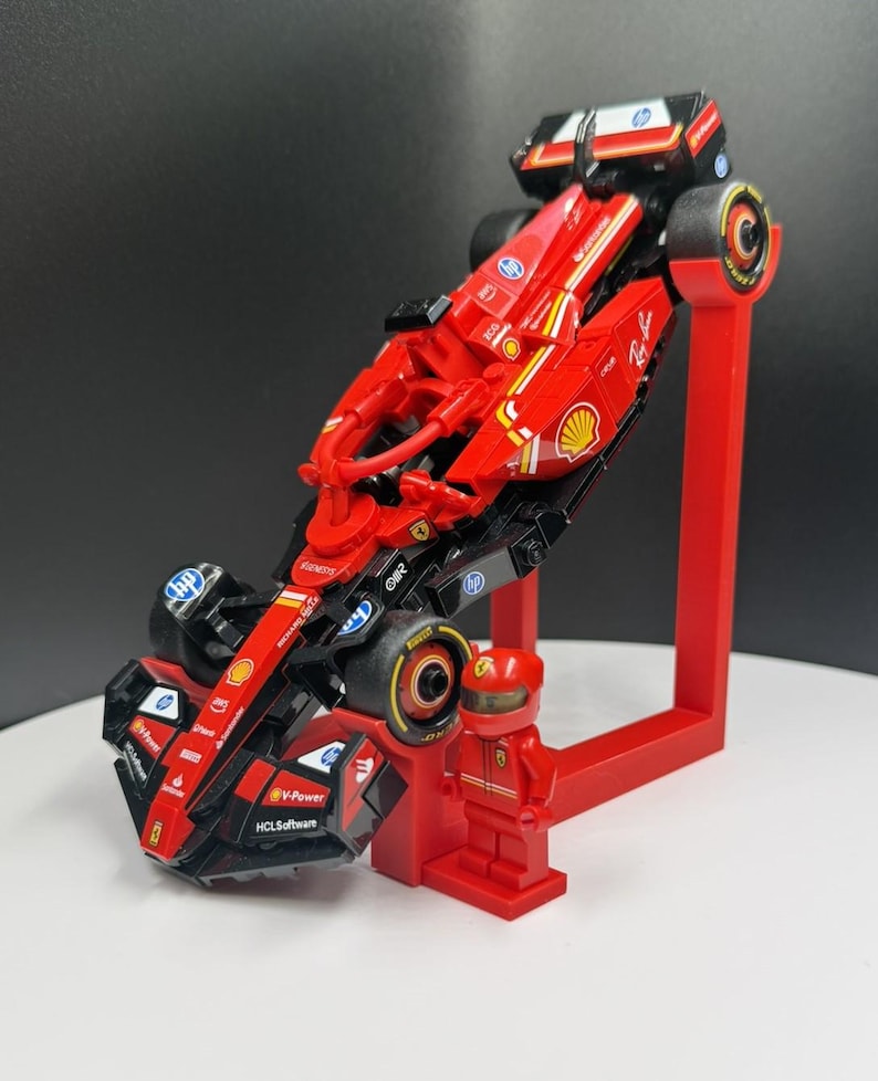 May include: A red and black Lego Formula 1 race car model with sponsor logos, displayed on a red stand. A red Lego figure of a driver stands next to the car. The car has black tires and yellow accents.