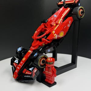 May include: A red and black toy Formula 1 race car with sponsor logos, displayed on a black stand. A red Lego figure in a racing suit and helmet stands next to the car. The car has yellow accents and black tires.
