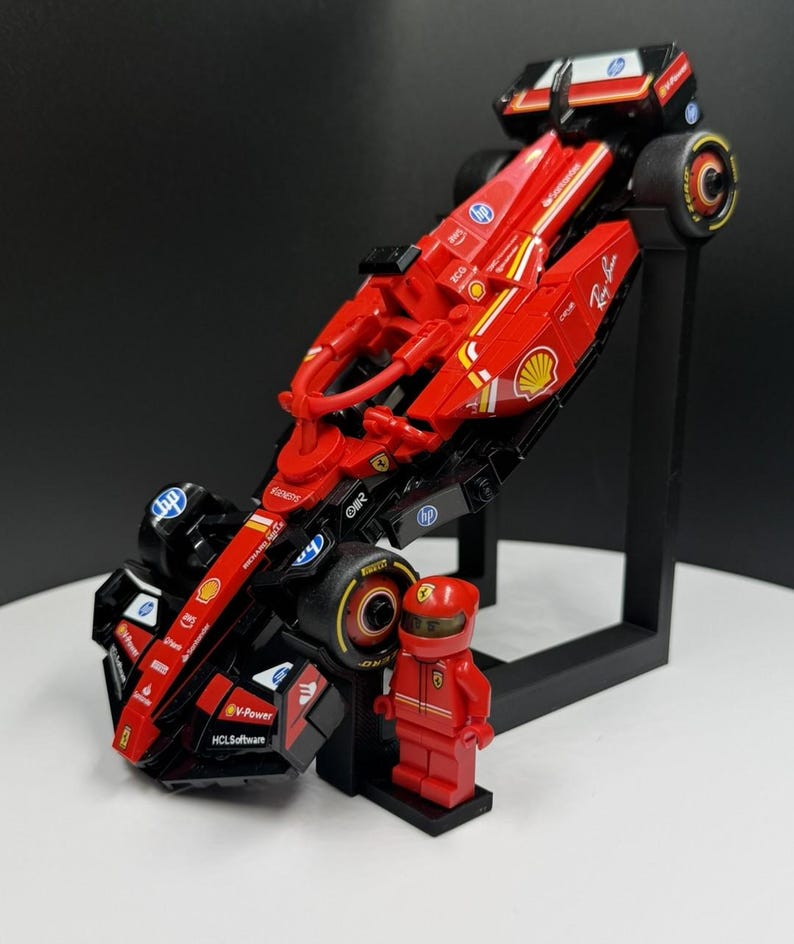 May include: A red and black toy Formula 1 race car with sponsor logos, displayed on a black stand. A red Lego figure in a racing suit and helmet stands next to the car. The car has yellow accents and black tires.