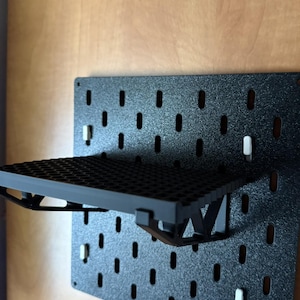 Peut inclure: Tableau perforé noir avec une étagère fixée. Le tableau comporte des trous espacés et une surface texturée. L'étagère est également noire et présente un motif en grille. L'étagère est soutenue par des supports et des chevilles blanches.