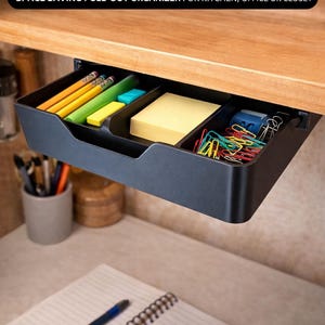 Pode incluir: Gaveta de armazenamento preta sob a prateleira com compartimentos, contendo lápis, notas adesivas e clipes de papel. A gaveta é montada sob uma prateleira de madeira. O texto na imagem diz: "Under Shelf Storage Drawer" e "SPACE SAVING PULL-OUT ORGANIZER FOR KITCHEN, OFFICE OR CLOSET."