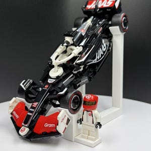 May include: A black and red Formula 1 race car model with the word "HAAS" in white. The model is displayed on a white stand, next to a matching driver figurine in a red helmet and white racing suit.