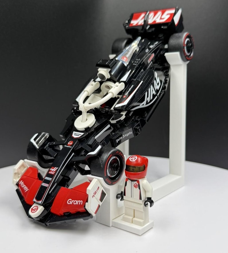 May include: A black and red Formula 1 race car model with the word "HAAS" in white. The model is displayed on a white stand, next to a matching driver figurine in a red helmet and white racing suit.